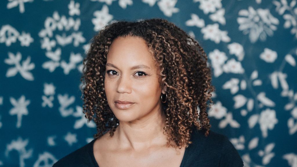 a woman posing looking at the camera in front of a floral wallpaper