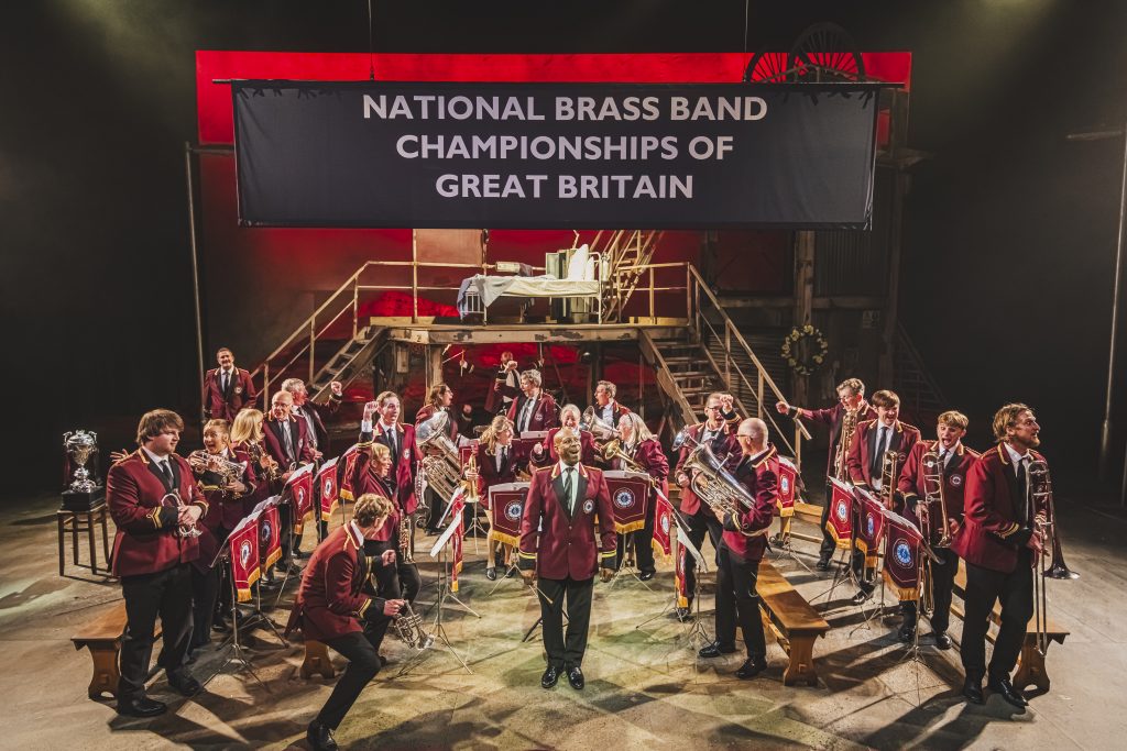 a theatre stage with an orchestral brass band stood together in matching red jackets 