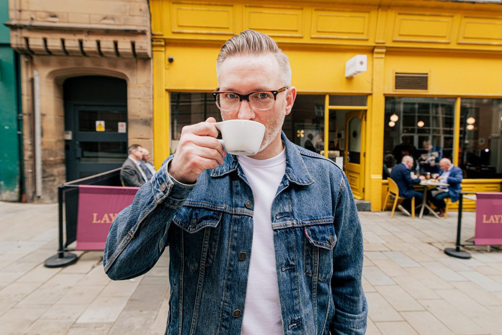 a person in front a bright yellow shop front, wearing a denim jacket and glasses and sitting from a small mug
