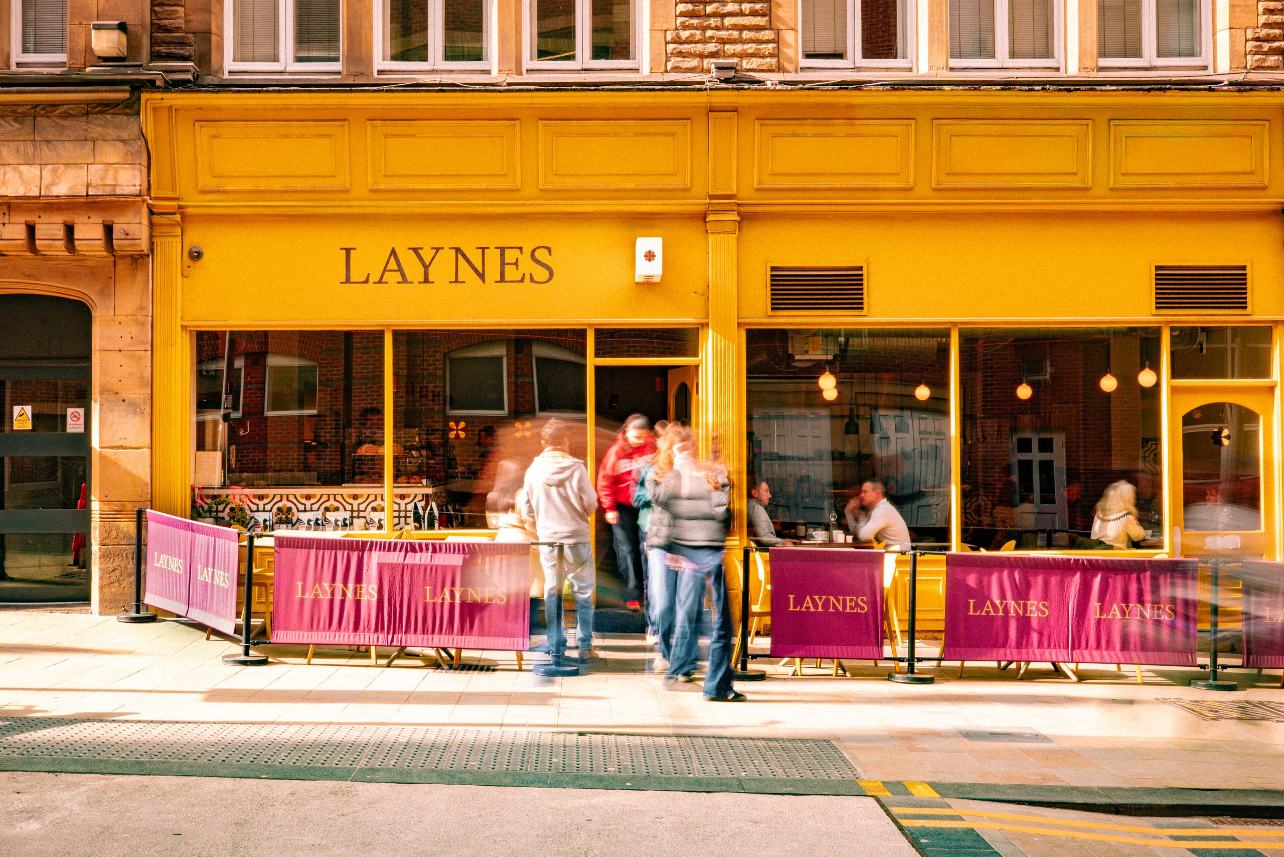 the front of a cafe with bright yellow windows, and people walking in and out of a doorway