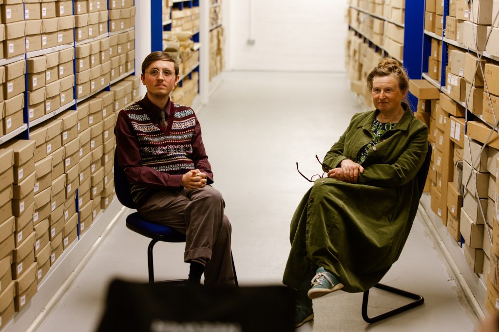 two people sat on chairs looking at the camera in a room filled with cardboard archival boxes