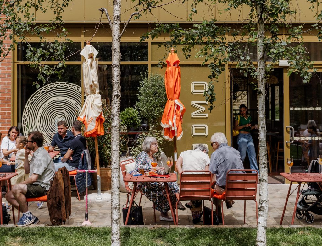 the outdoor terrace of a restaurant with people sat outside in the sun on wooden tables and chairs