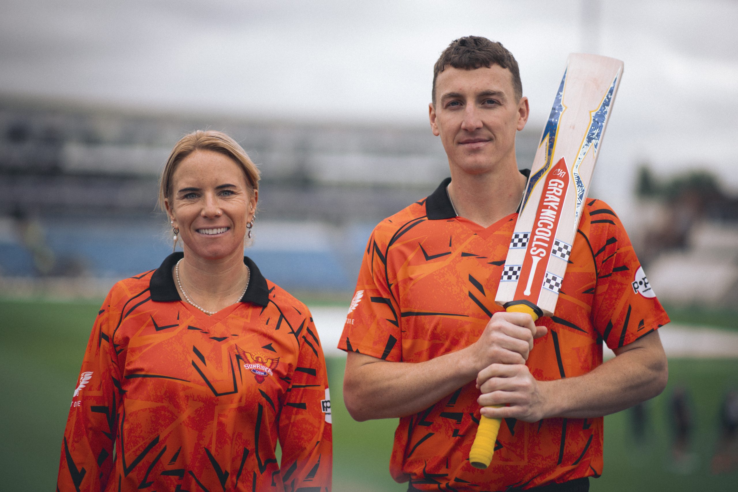 two people stood in a field, one holding a cricket bat, both in orange sports tops