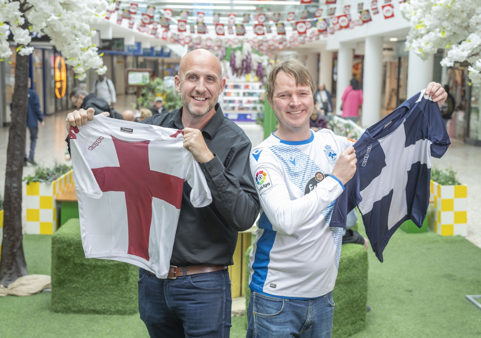 two people stood inside a shopping centre decorating with flags whilst holding a tshirt each, one with the england flag and the other scottish flag