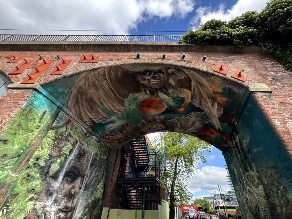a mural underneath a bridge featuring botanical and underwater patterns, a hand with a tribal pattern and two owls with a moon inbetween