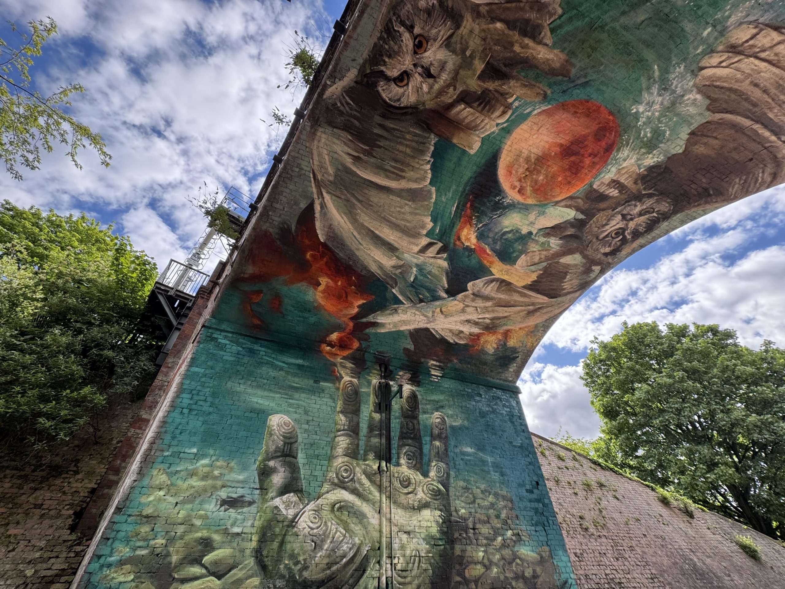 a mural underneath a bridge featuring botanical and underwater patterns, a hand with a tribal pattern and two owls with a moon inbetween