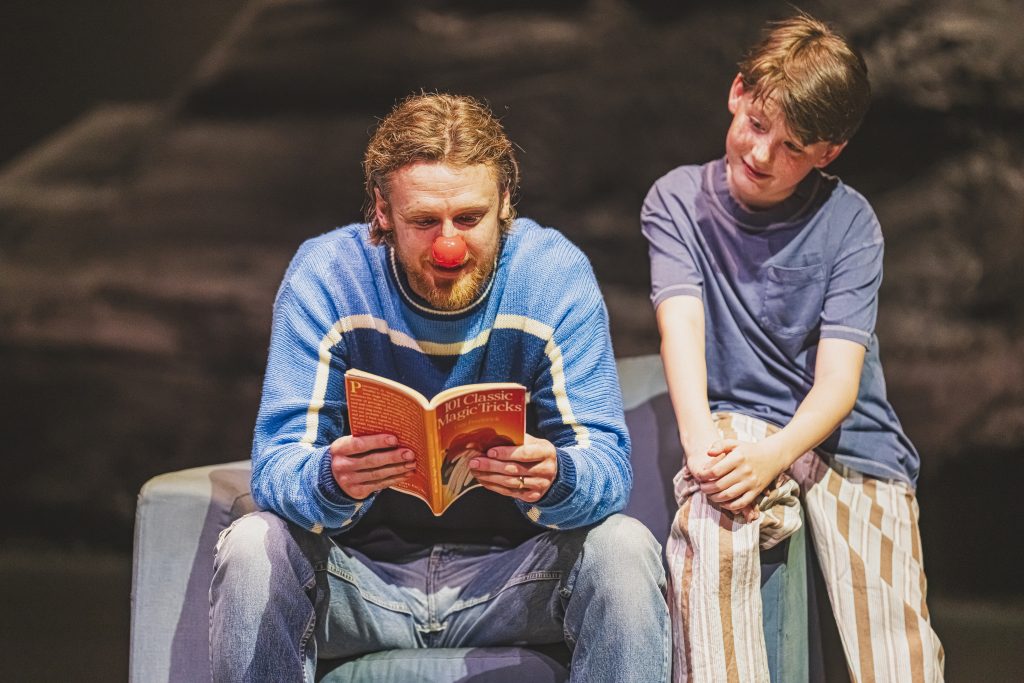 a theatre stage, with a man sat in a chair reading a book and a young child sat on the arm of the chair also reading