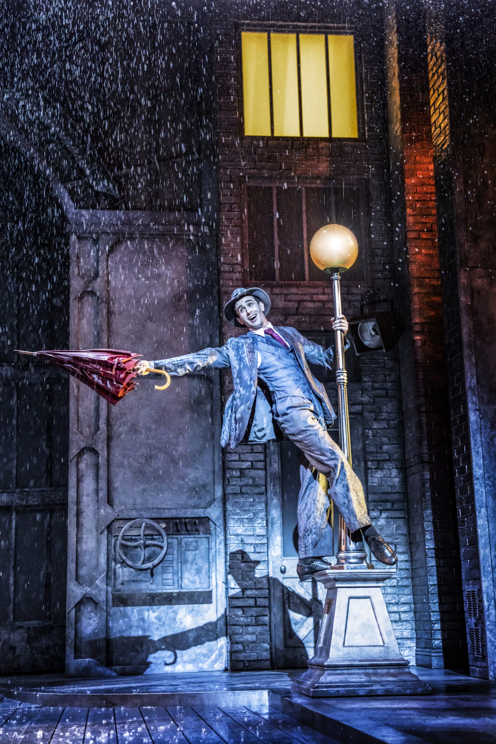 a person performing on stage in a suit holding an umbrella whilst hanging off a lamp post with a raining backdrop