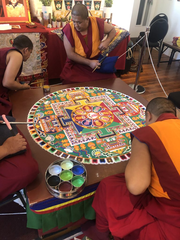 buddhist monks in orange and red robes doing a mandala on a table, a decorative patterned and colourful picture made with sand