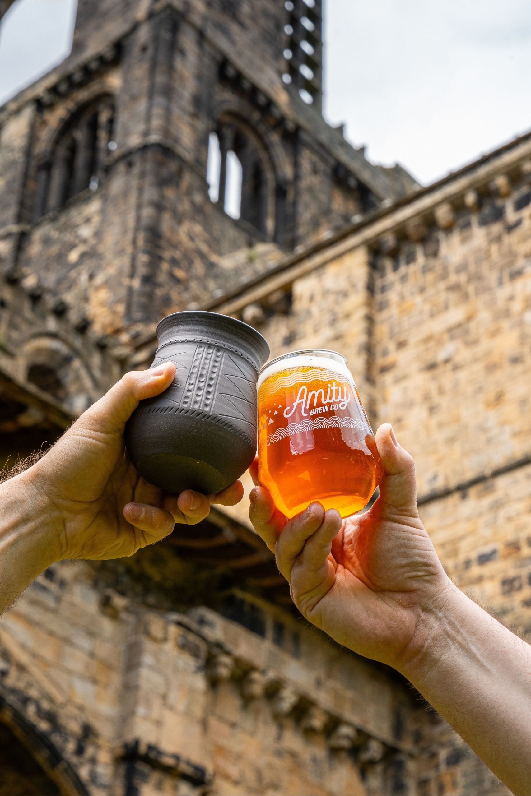 two hands holding a glass with an amber liquid and a black cup in front of the ruins of an old abbey