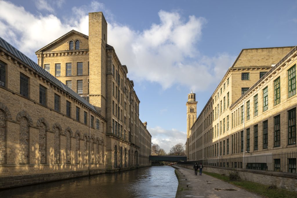 Exterior view of Salt Mills in Saltaire with canal running through sunlit buildings beneath blue skies