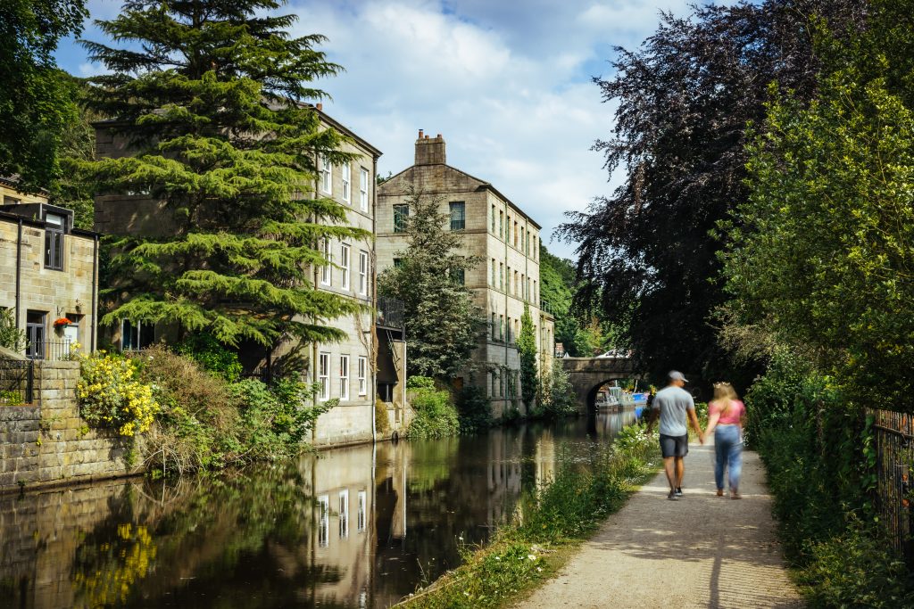 Two people walk hand in hand away from the camera along the canal in a sunny Hebden Bridge