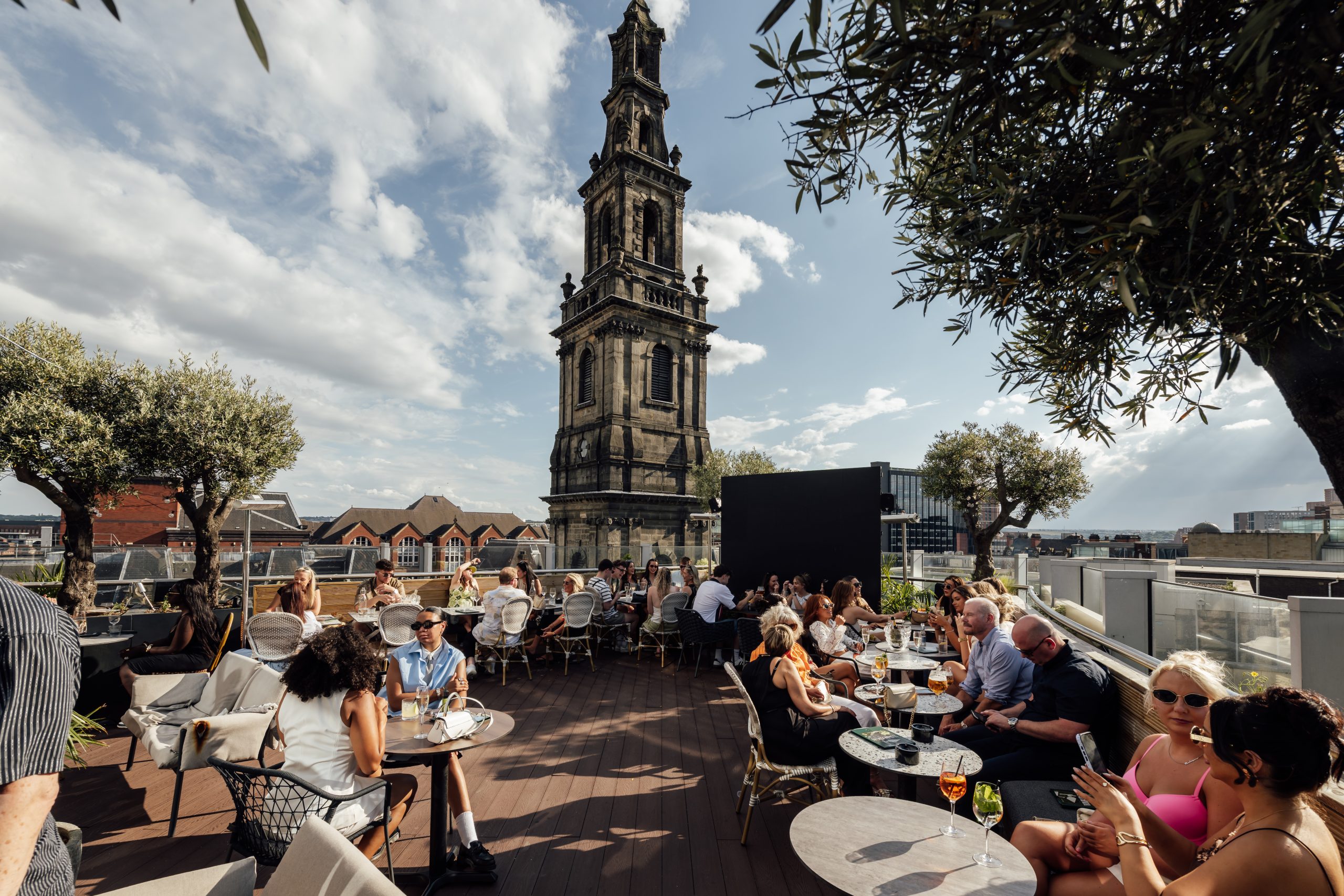 the rooftop garden of a restaurant with a church spire behind, and people sat at round tbales eating and drinking