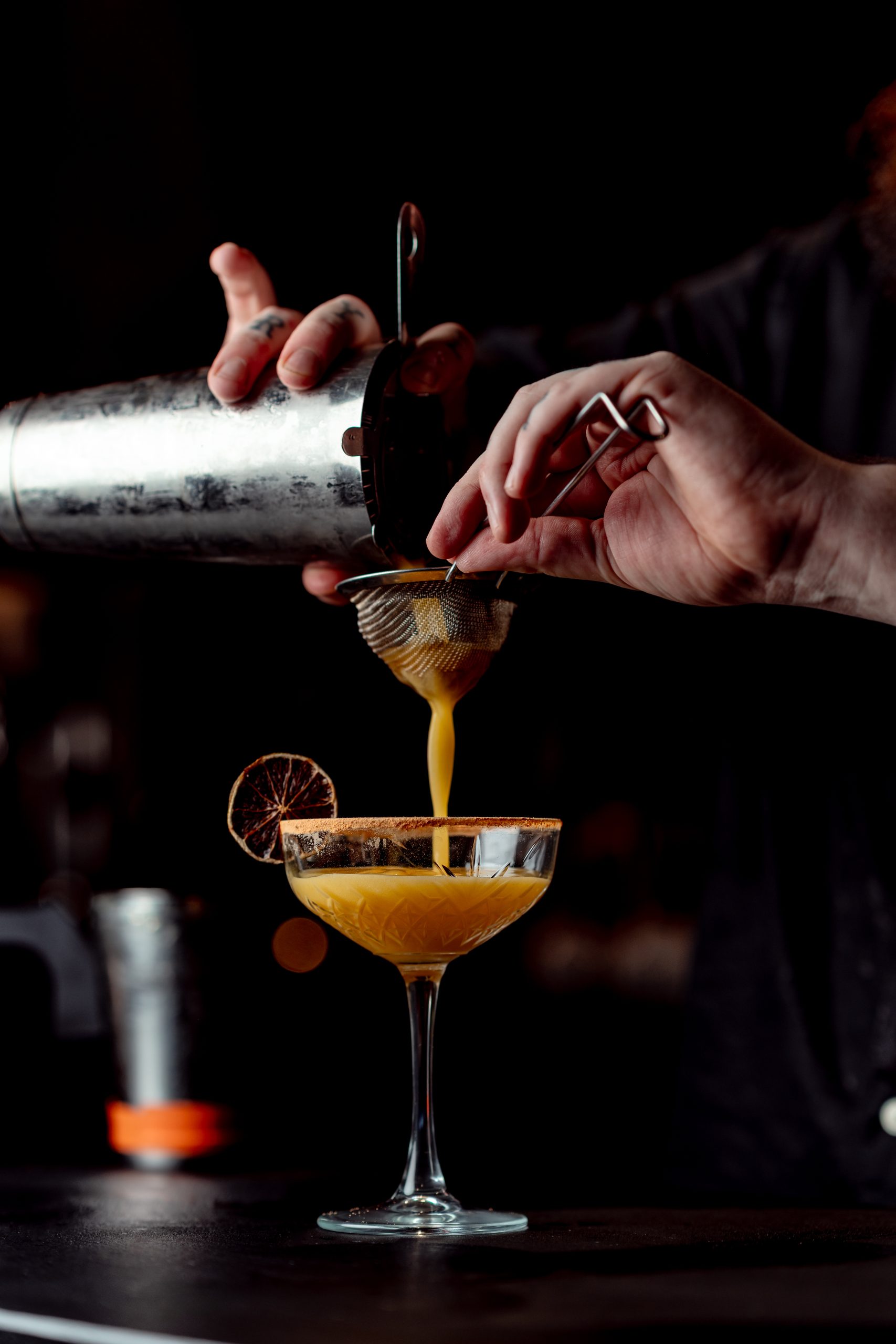 two hands pouring form a cocktail shaker through a sieve into a cocktail glass
