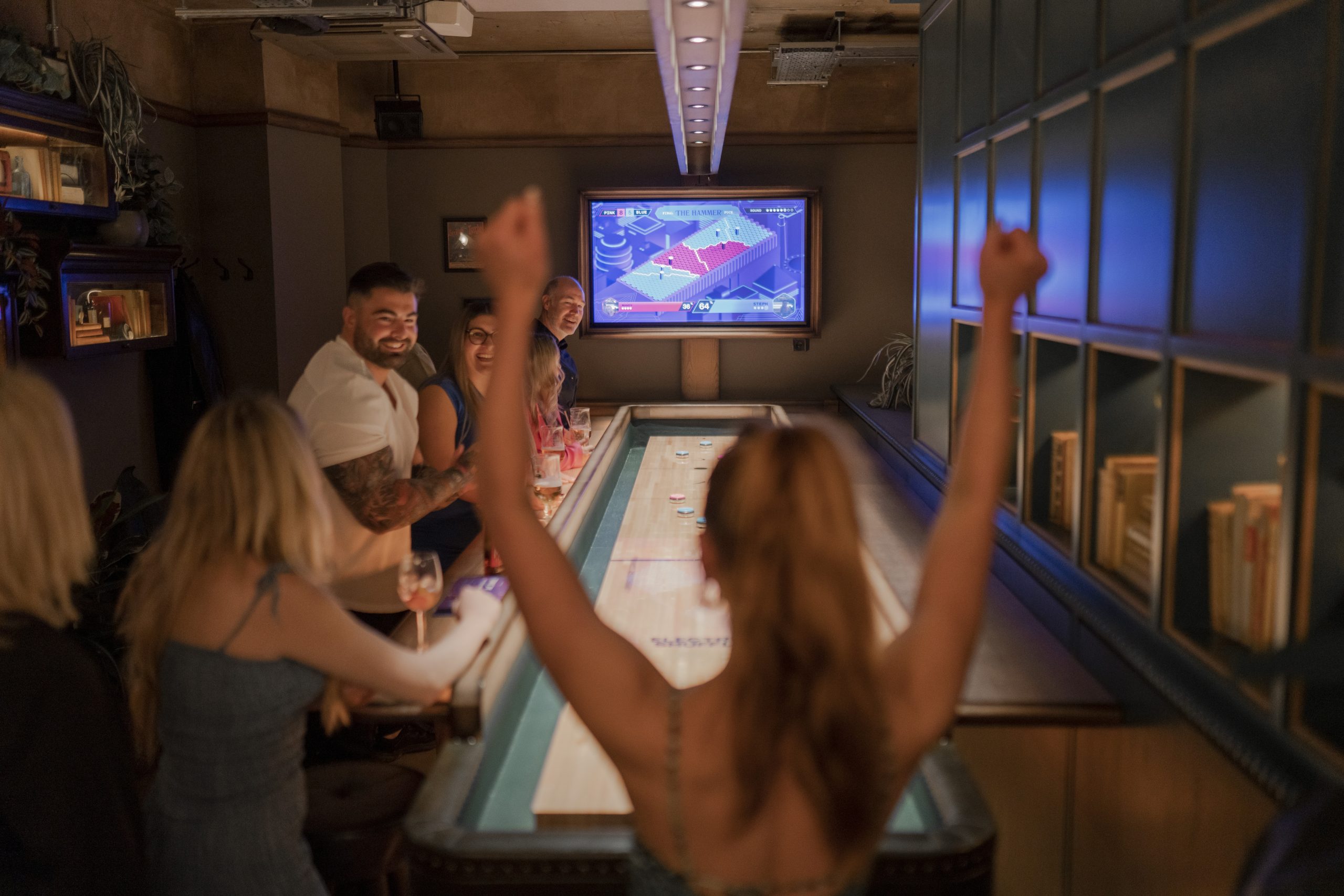 a small group of people stood around an illuminated shuffle board table with a large tv screen on the opposite side