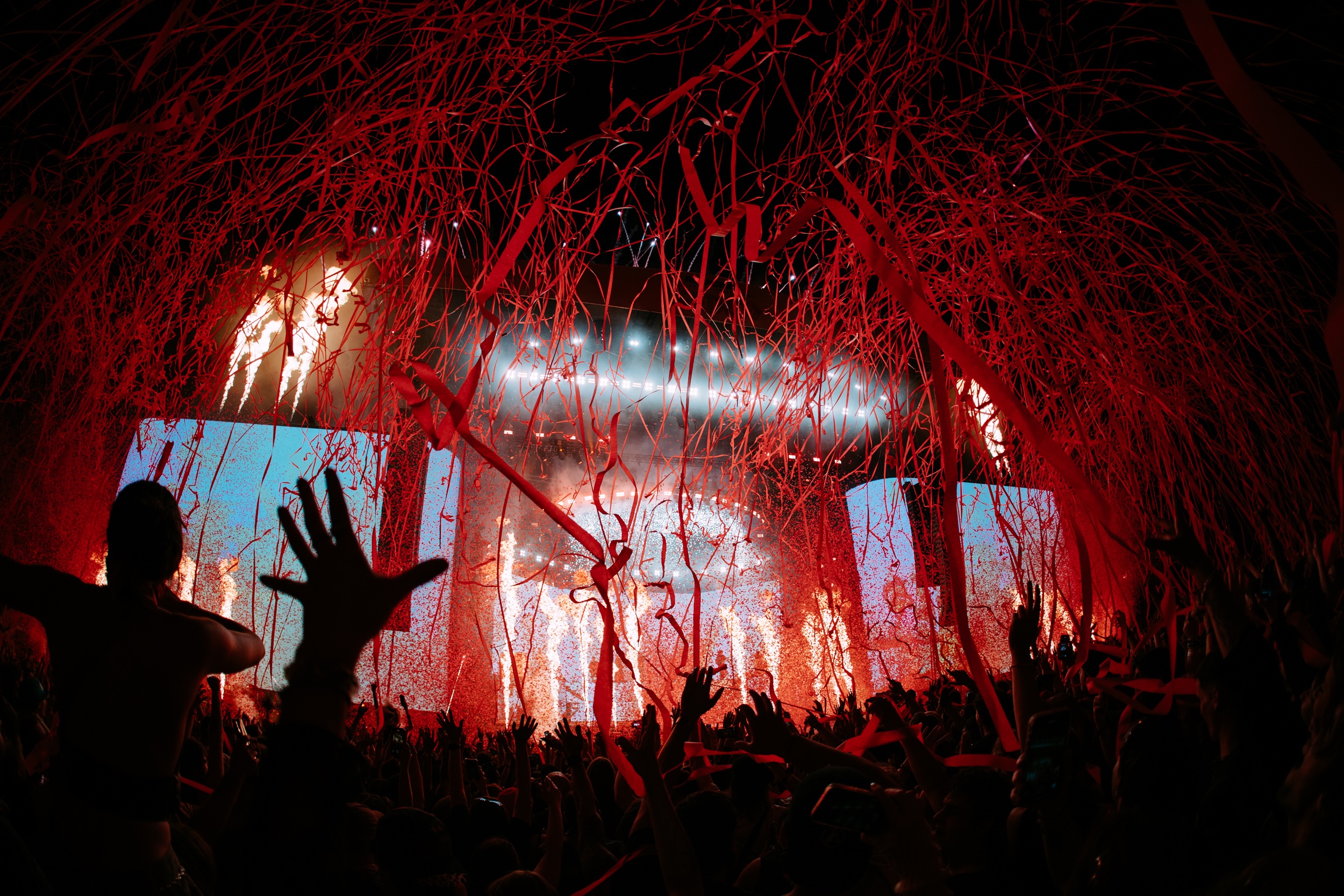 a large outdoor stage at a music festival at night, with a crowd of people with their arms up and bright lights and confetti from the stage