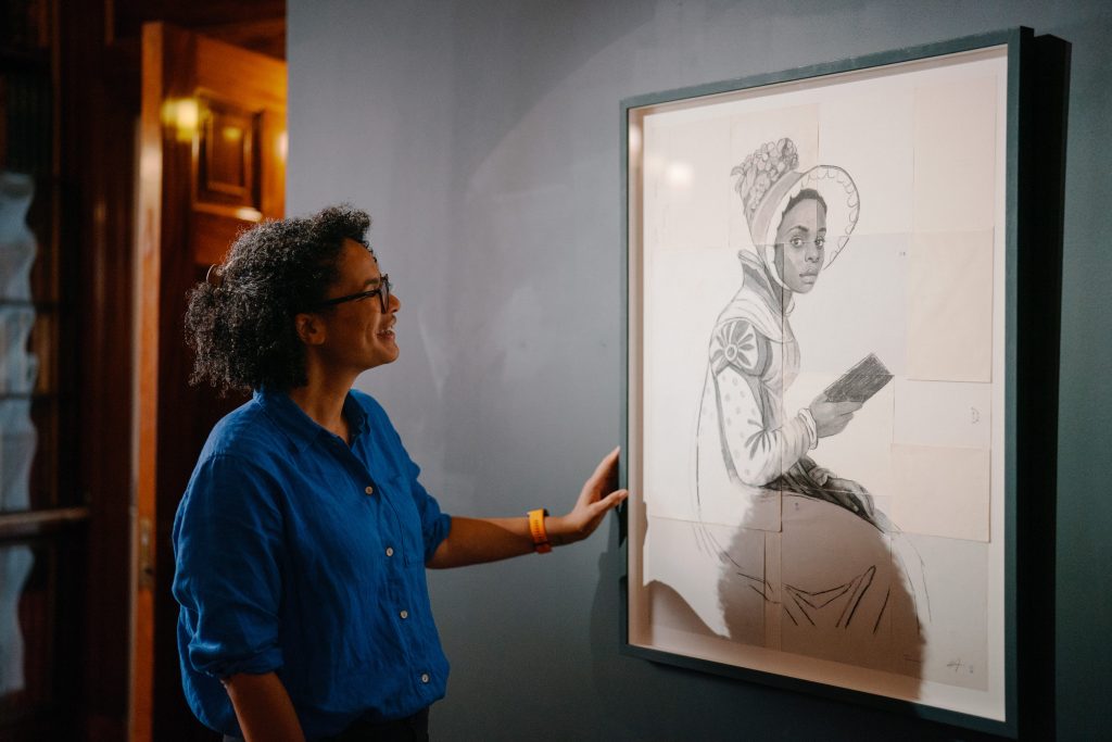 a woman looking at an artwork on a wall, of a young woman in regency dress and a bonnet reading a small book