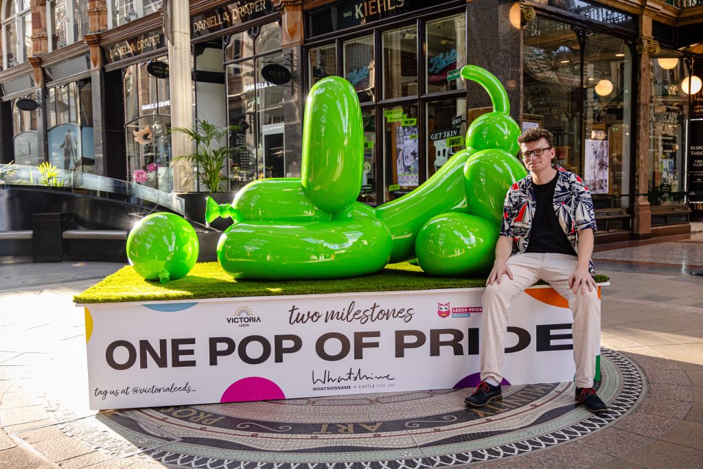 a large sculpture of a balloon dog in green on a stand saying one pop of pride and a person sat on the stand, sitting in the middle of a shopping centre with old victorian architecture and a stained glass roof