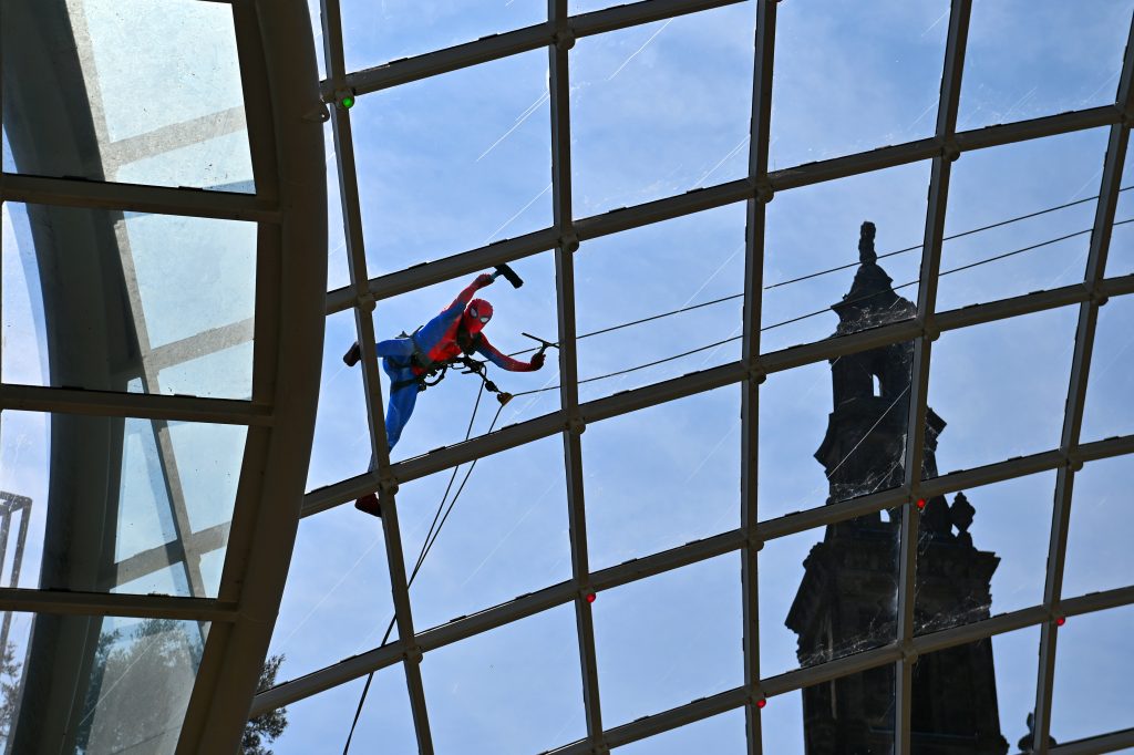 a person on the roof of a glass dome dressed in a spiderman costume with a church spire visible behind