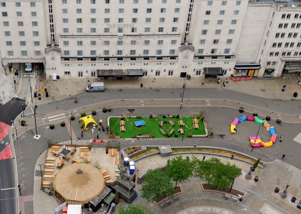an aerial view of a square in between lots of tall buildings with tents, greenery and colourful benches