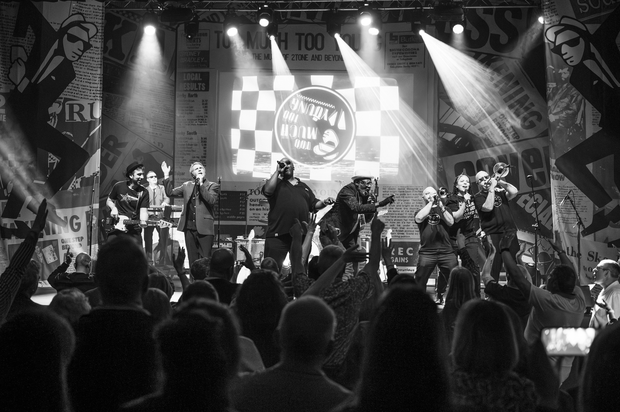 a group of musicians on a stage including singer, guitarist, trombone and trumpet players, in front of a crowd of people