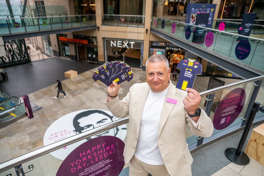 a man holding cards on the second floor of a shopping centre looking at the camera, with a graphic of a man's face on the floor below