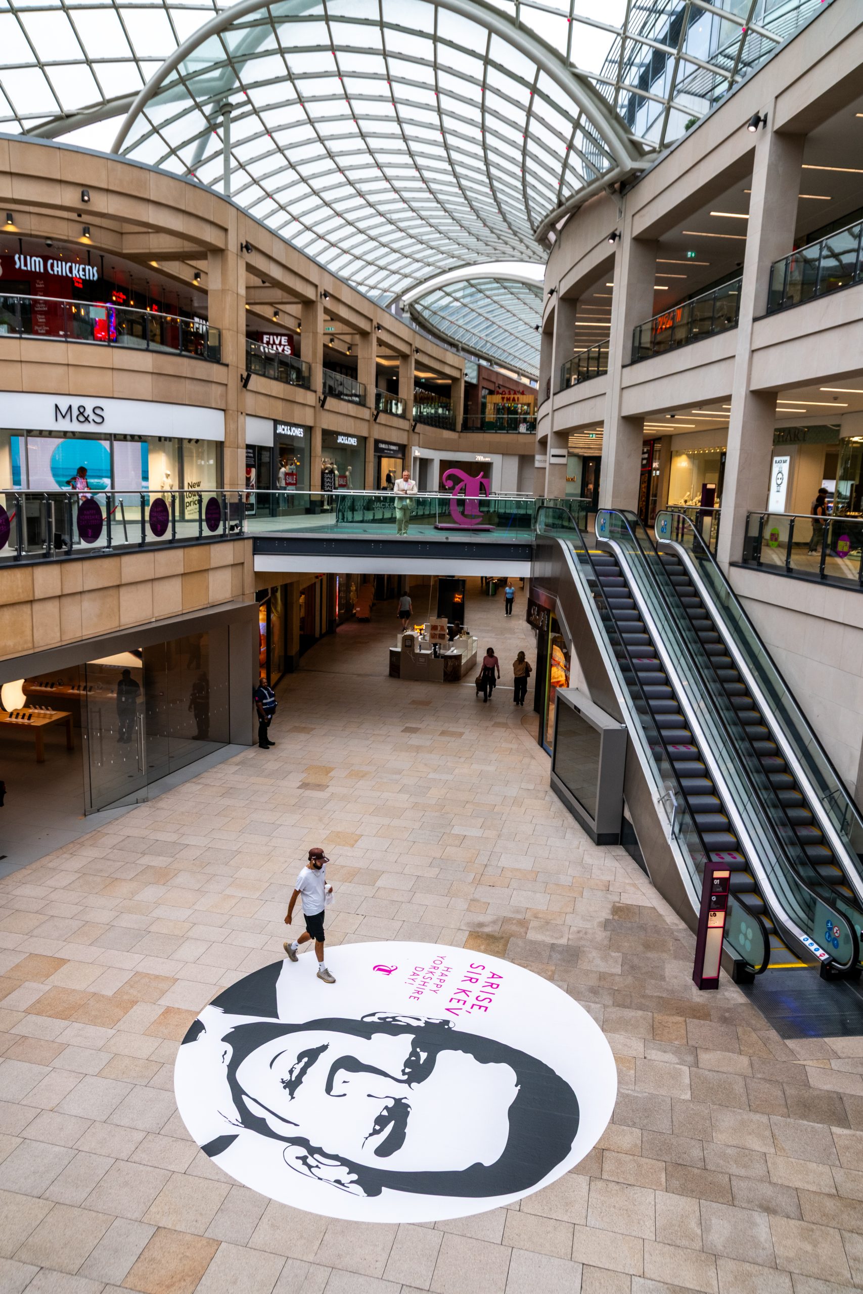 the interior of a shopping centre with three floors, a large glass roof, and a picture of a man on the ground floor