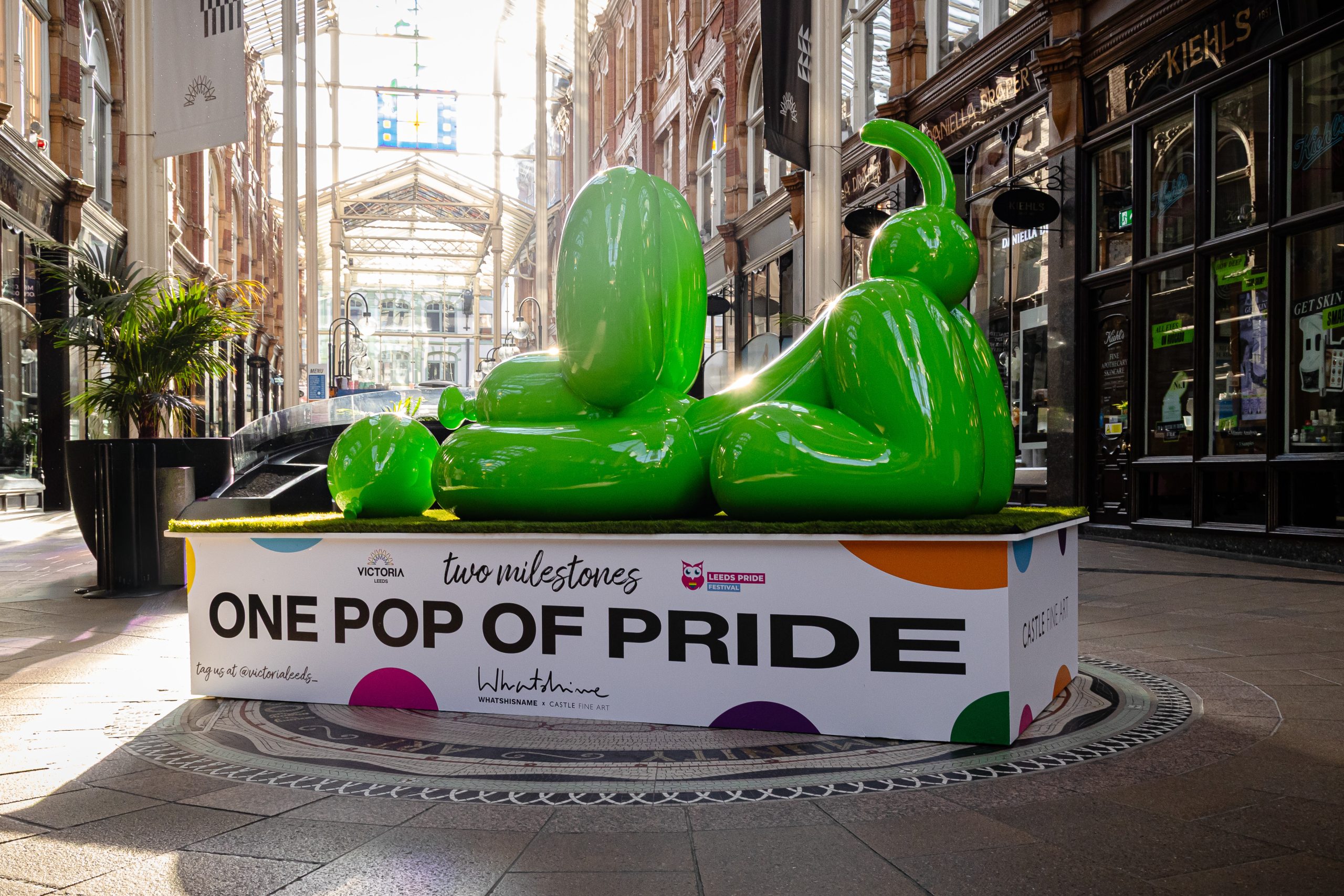 a large sculpture of a balloon dog in green on a stand saying one pop of pride, sitting in the middle of a shopping centre with old victorian architecture and a stained glass roof