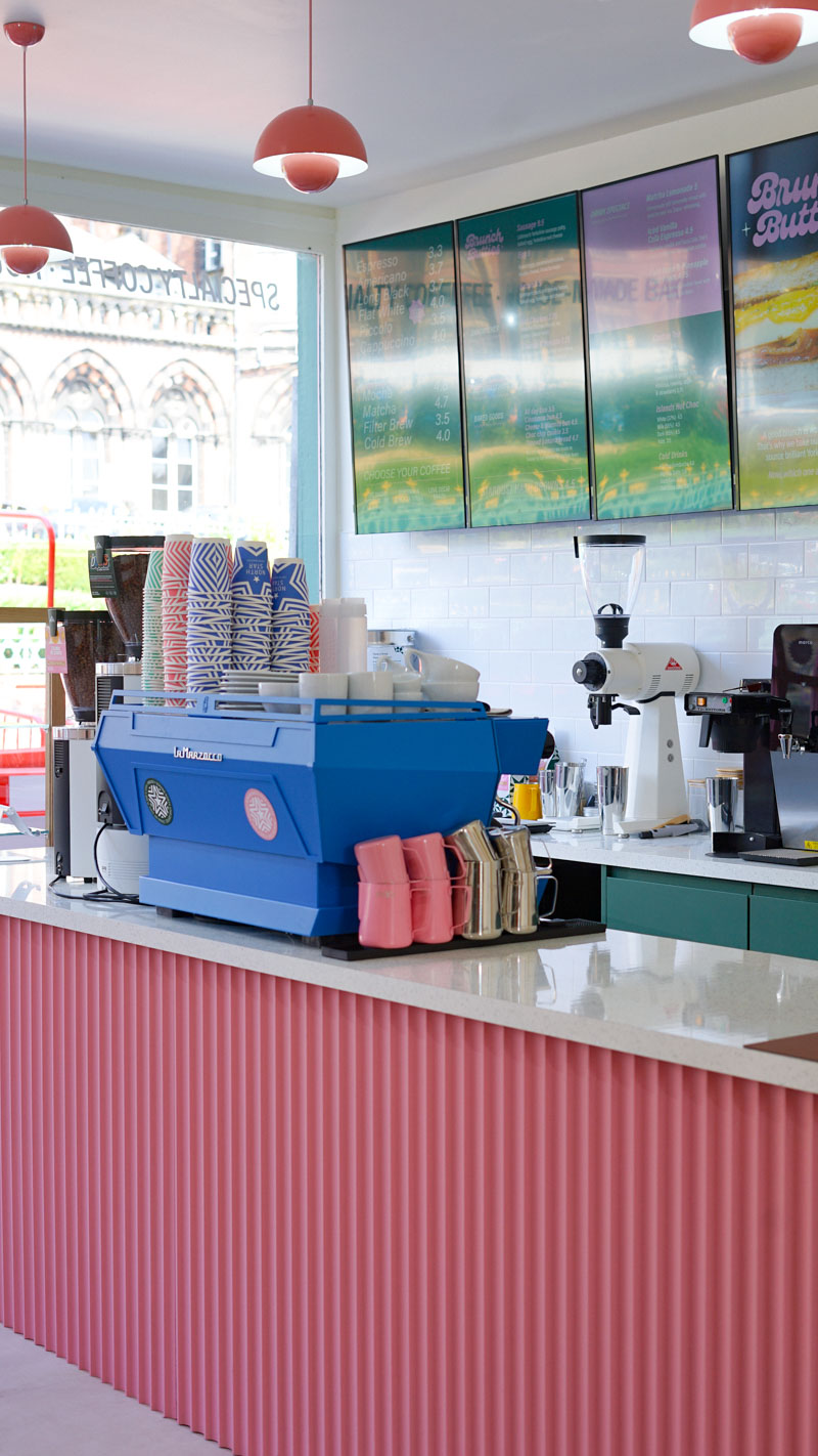 the interior of a coffee shop, with a large window front letting in natural light, a bright blue espresso machine, pink counter and screens on the wall above
