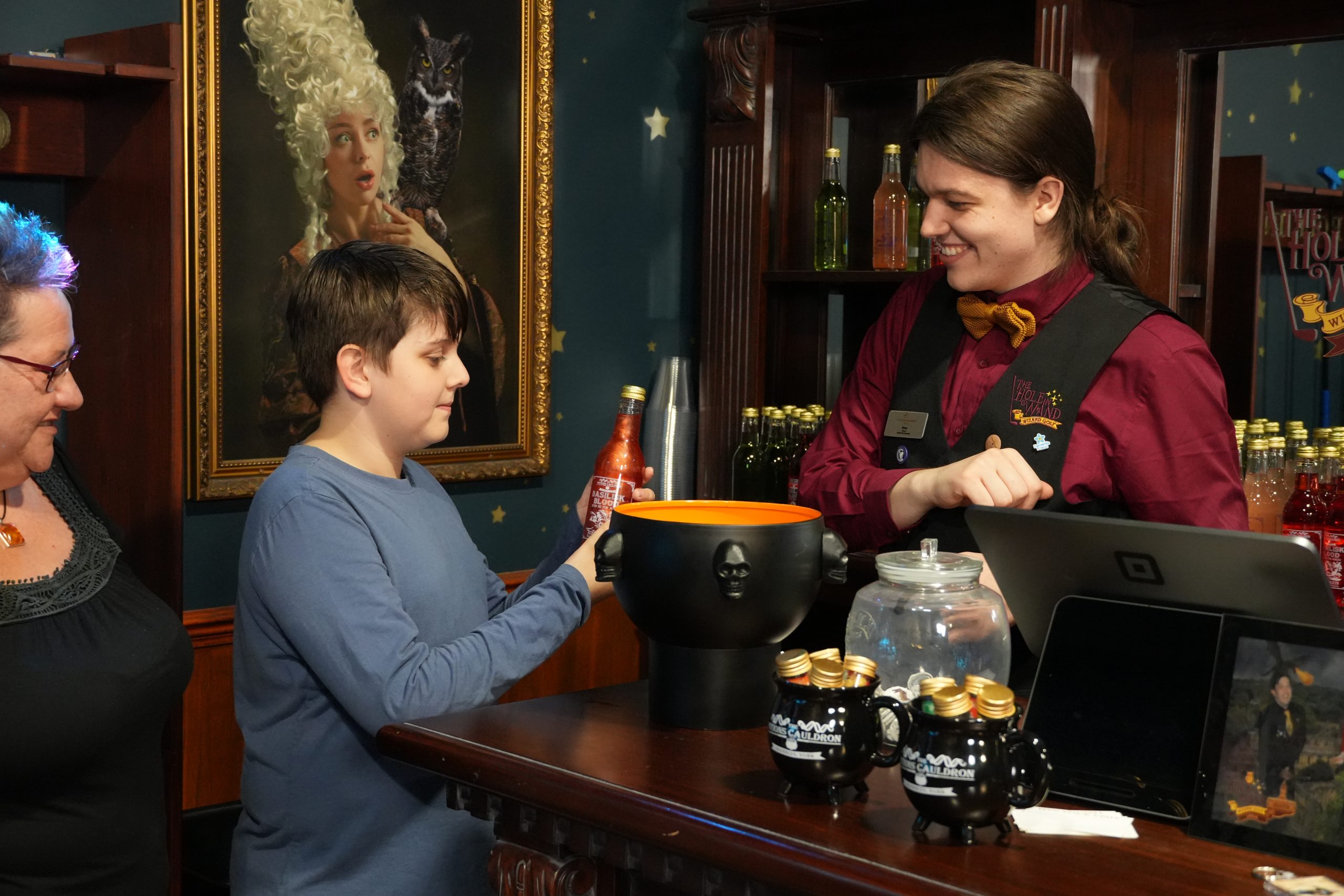 a person stood behind a desk, with a cauldron in front of them handing a glittery bottle to a young boy
