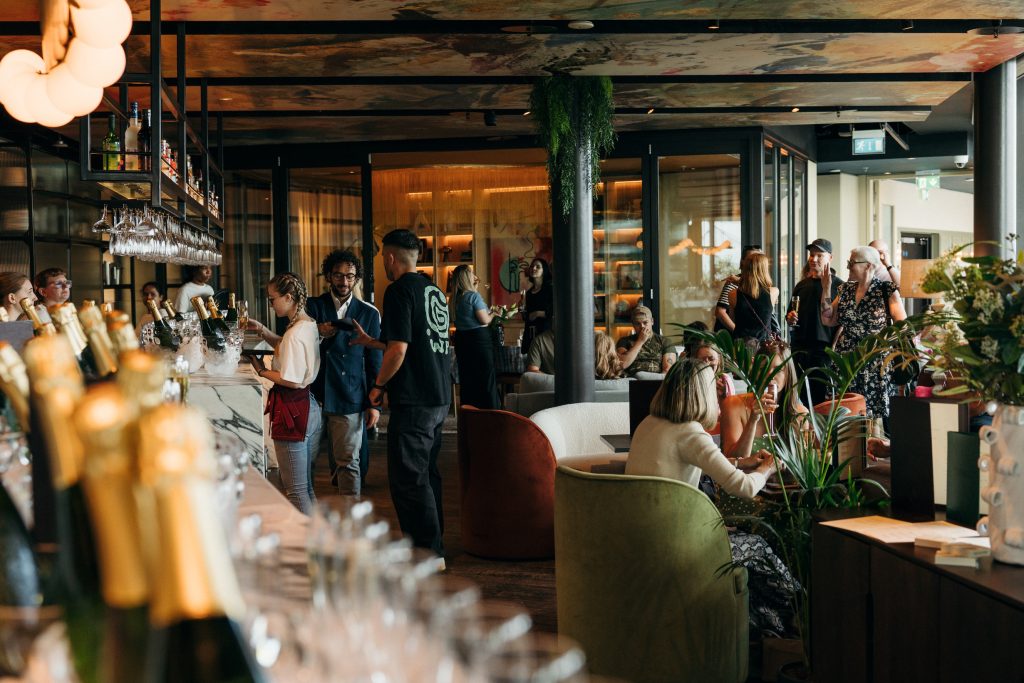 View of a crowd in CrowdedHouse enjoying a sit-down dinner with rows of champagne bottles in foreground