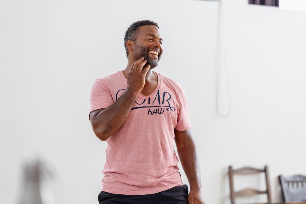 a man stood in a white room, with a wooden chair behind him