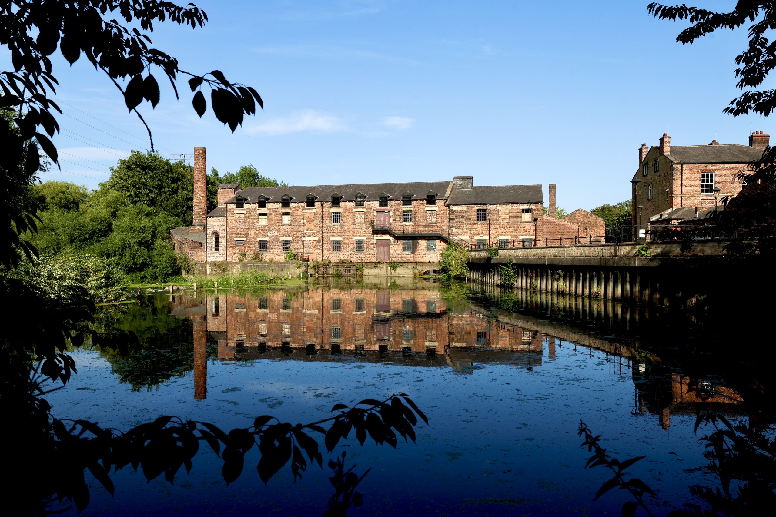 a large old red brick mill in front of a river reflected in the water