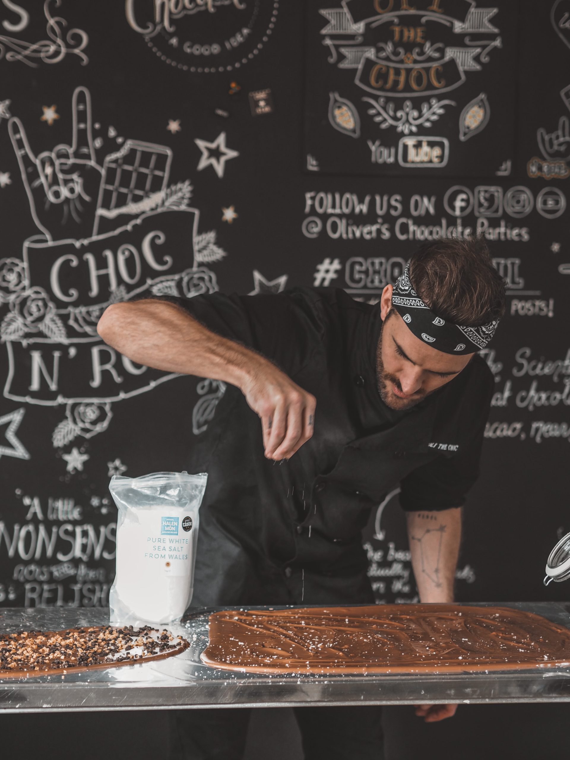 a person in a chef coat and bandana sprinkling salt onto a slab of melted chocolate in front of a blackboard with white chalk patterns