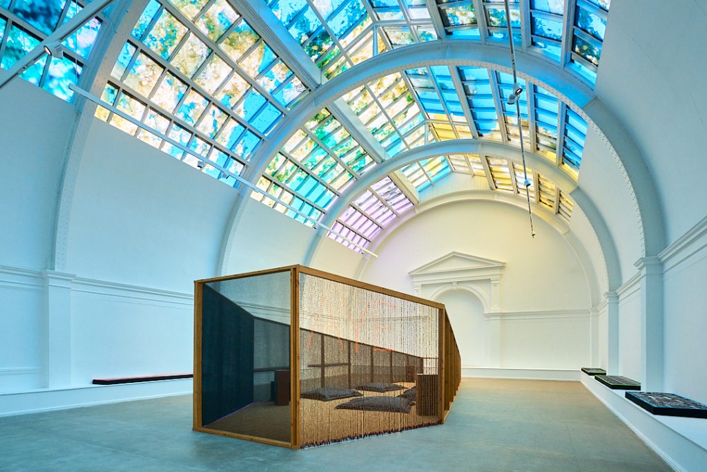a large room with a colourful glass window ceiling and a wooden frame structure in the middle of the floor with netting walls