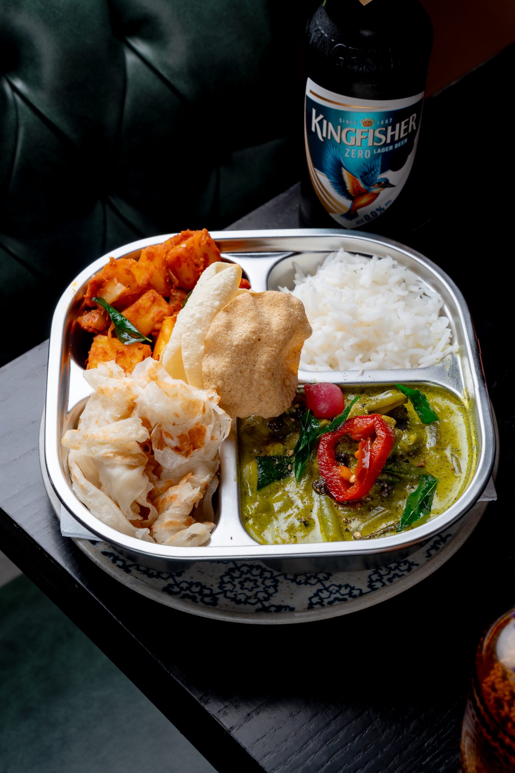 a silver tray with four compartments, one with rice, and the other three with colourful food, with a kingfisher beer bottle next to it