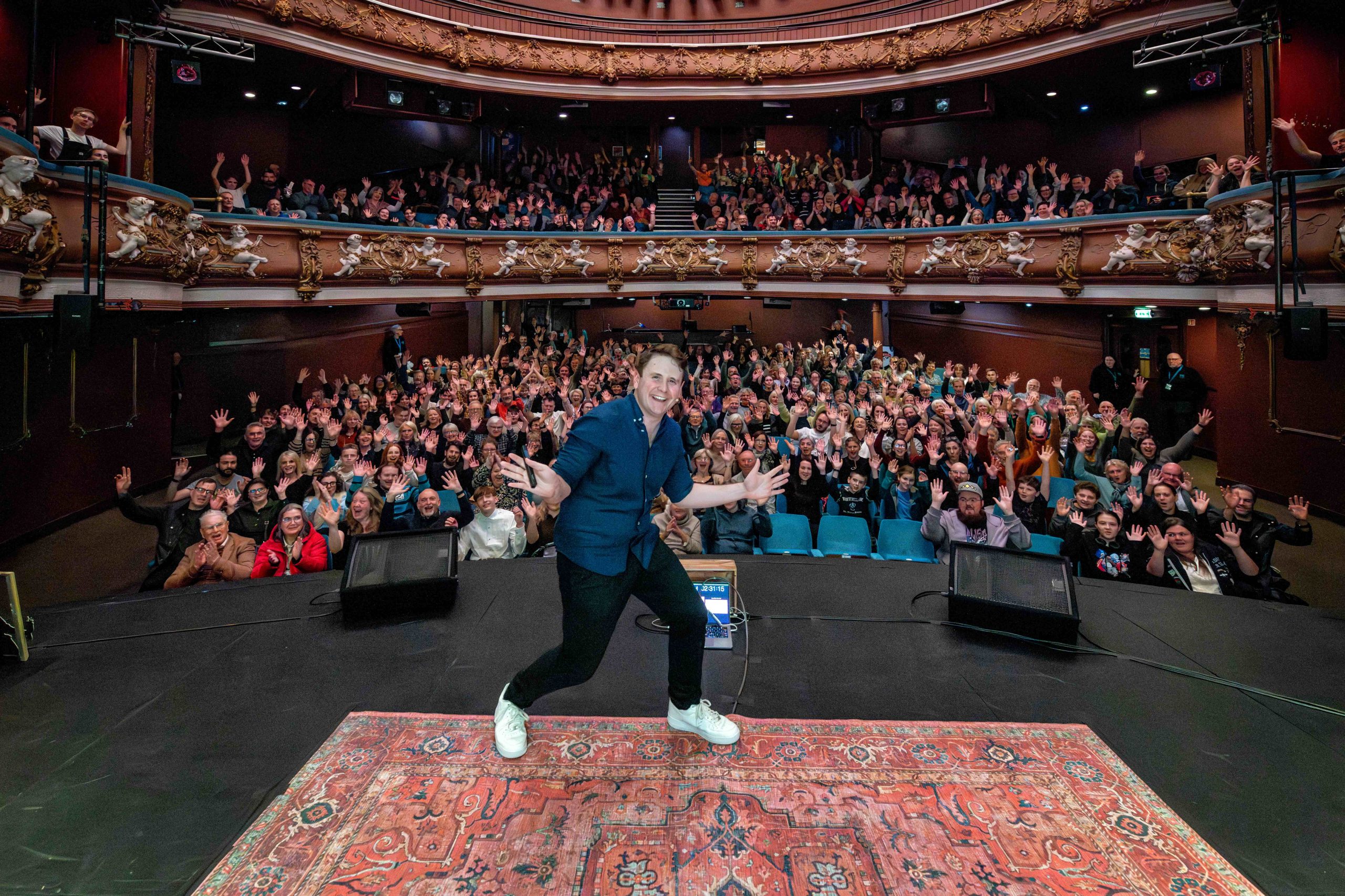 a person on a stage at a theatre facing the camera with the crowd behind him