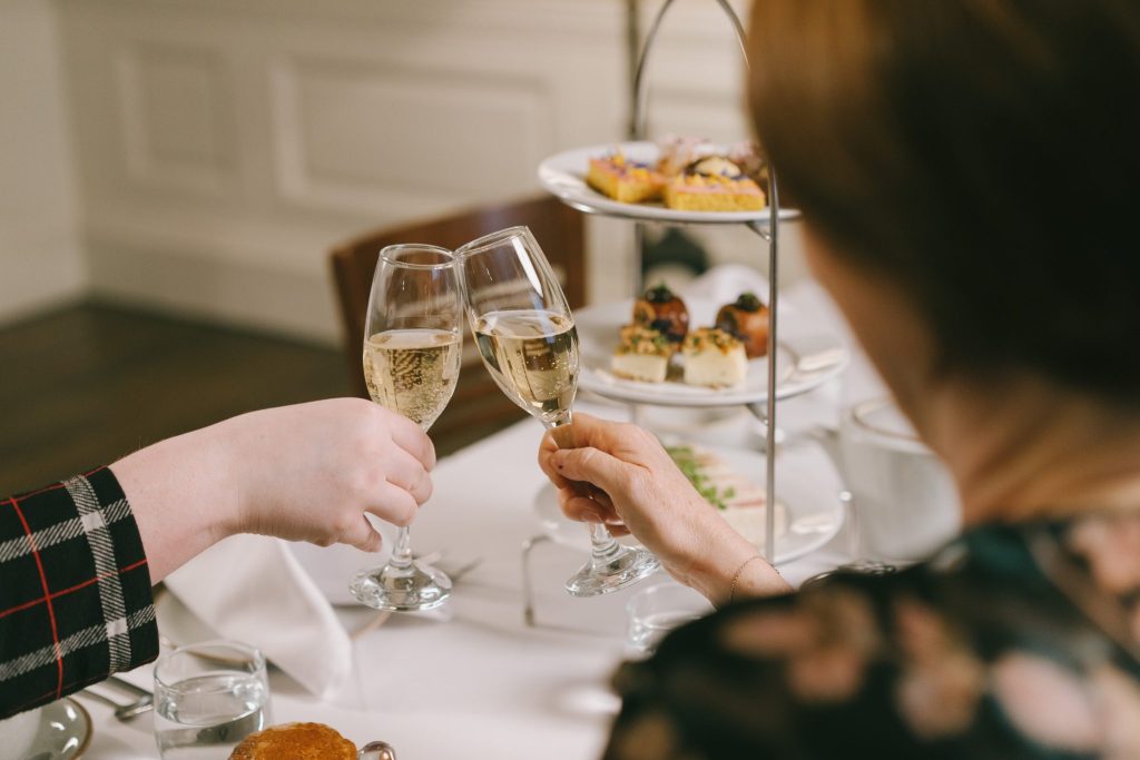 an over the shoulder view of an afternoon tea with a three tiered plate of food and the people clinking two flutes of champagne together
