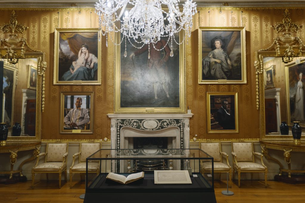 a room with a large chandelier, golden framed paintings on the wall and a glass cabinet on the floor with two books inside