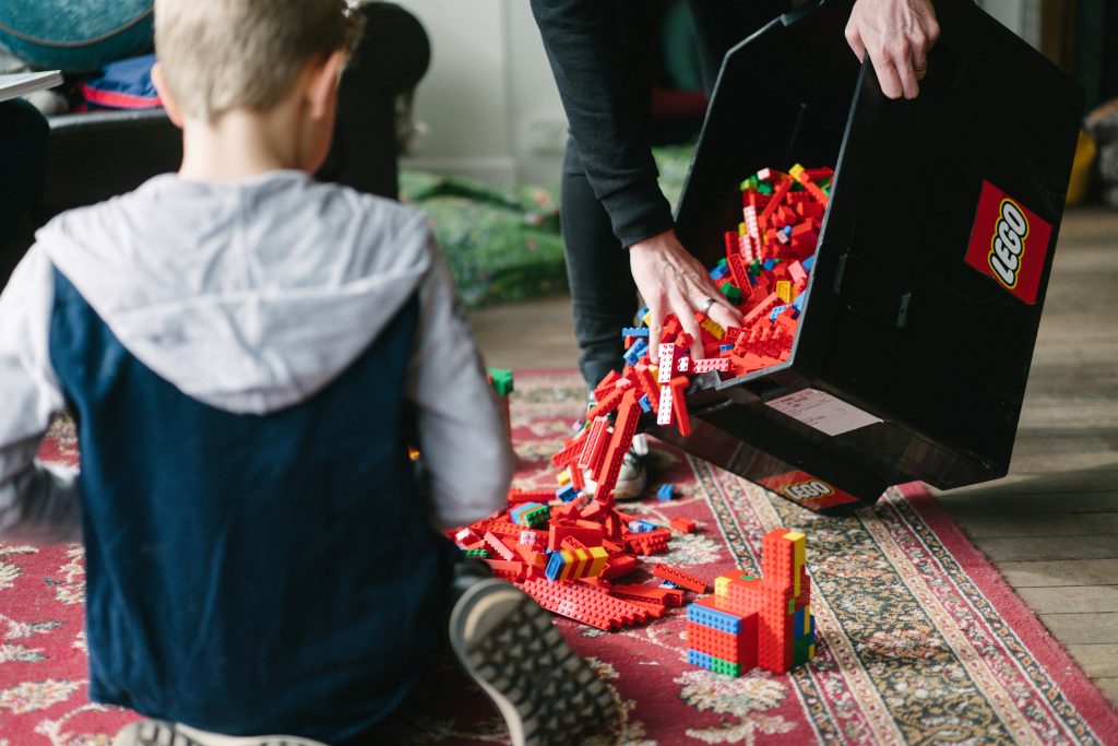 a child sitting on a floor inside a room with an adult pouring out lego pieces from a plastic tub