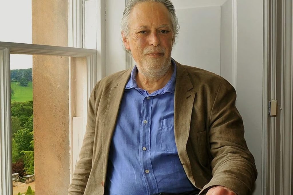 a portrait photo of a man in a blue shirt and light brown jacket sat at a window
