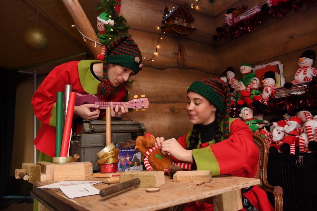 two people dressed as elves in red and green outfits inside a wooden room with colourful toys surrounding them