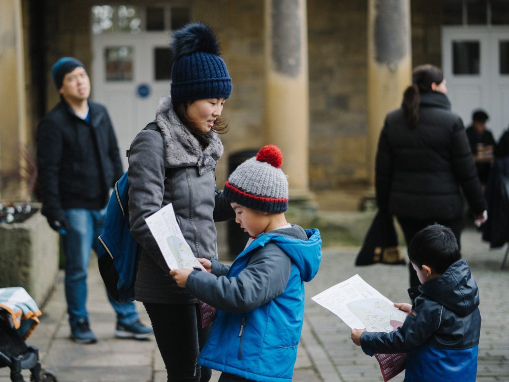 a stone courtyard building with columns and a roof around the edge, people walking around a small child in the foreground looking at a piece of paper