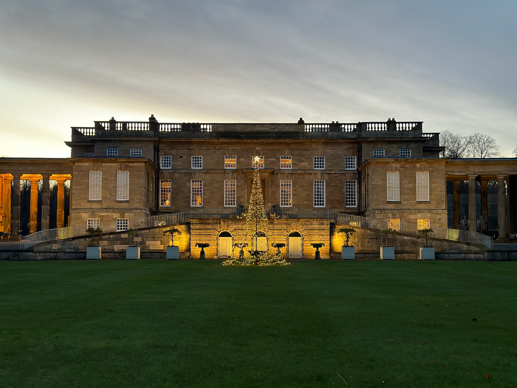 the front of a large historic home at dusk with a glowing christmas tree outside