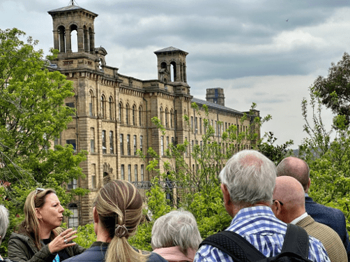 a group of people with someone facing them gesturing to a large mill complex behind them