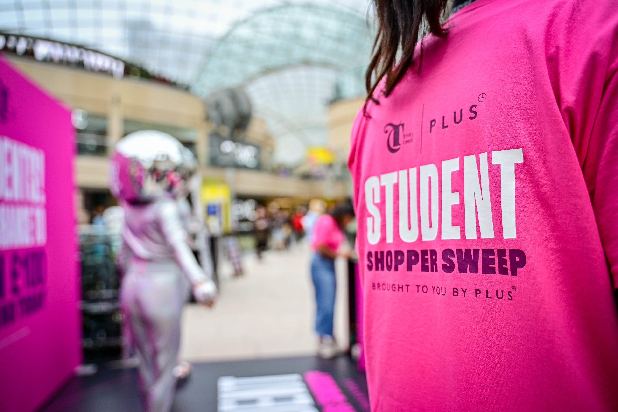 a person stood close to the camera with a tshirt on advertising a student shopping event with a crowd behind in a glass roofed building