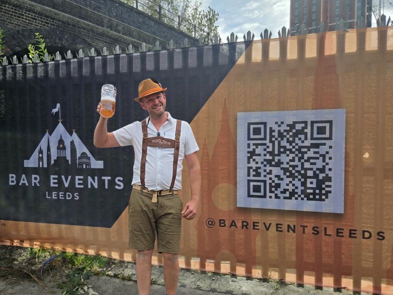 a man in traditional German lederhosen stood in front of a wall advertising bar events uk