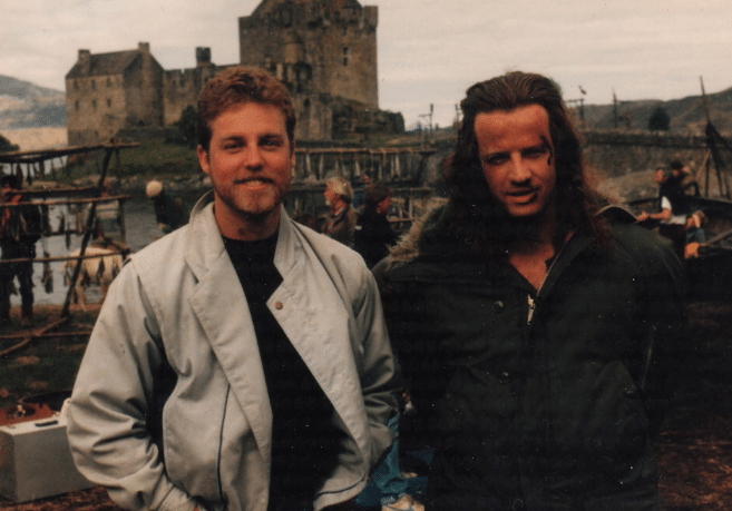two men stood together in front of a castle, one in a grey jacket and another with fake injuries and a large black coat