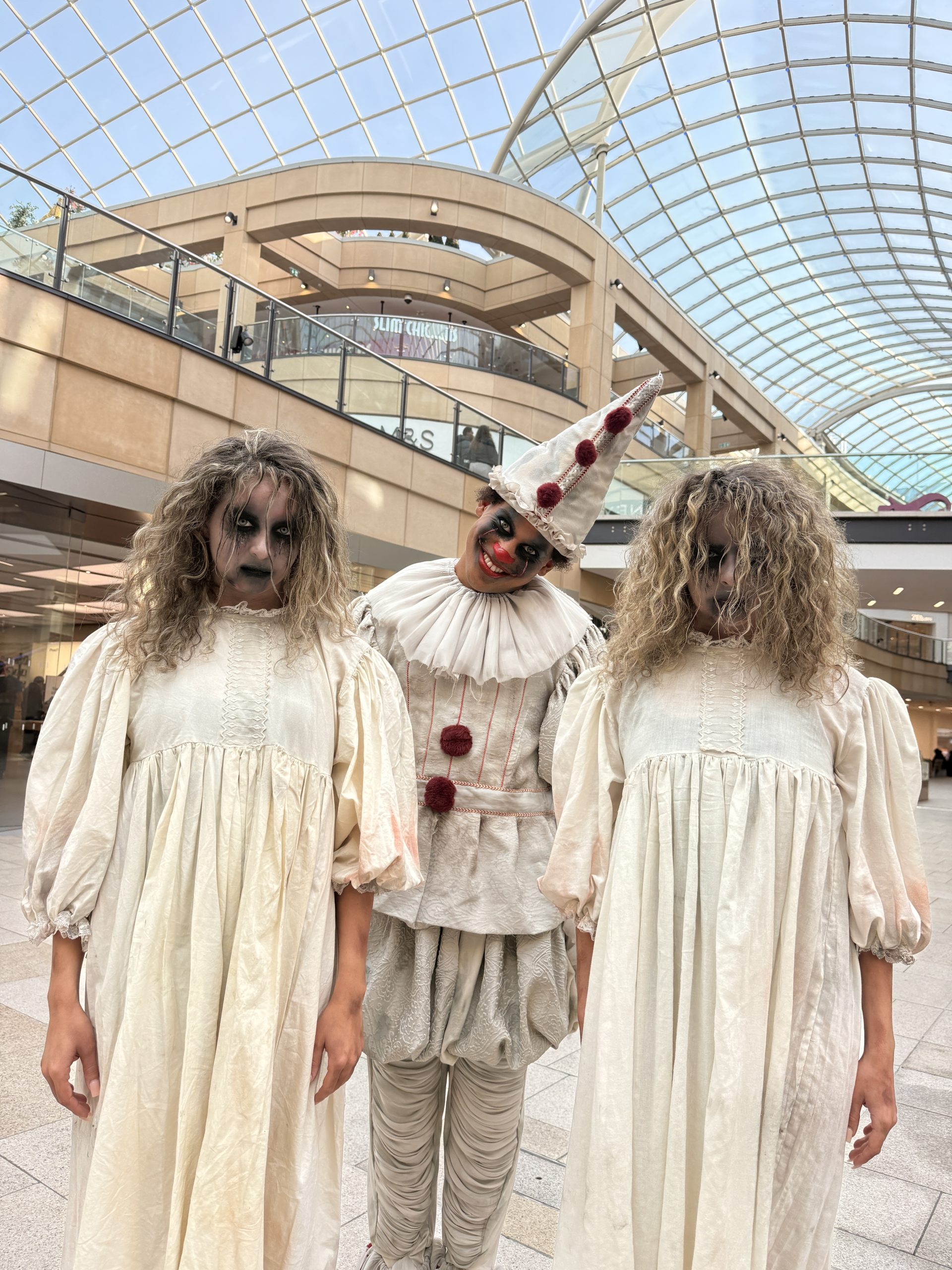 three people inside a glass roofed building, one dressed as a scary clown and the other two as scary twins with dark makeup on