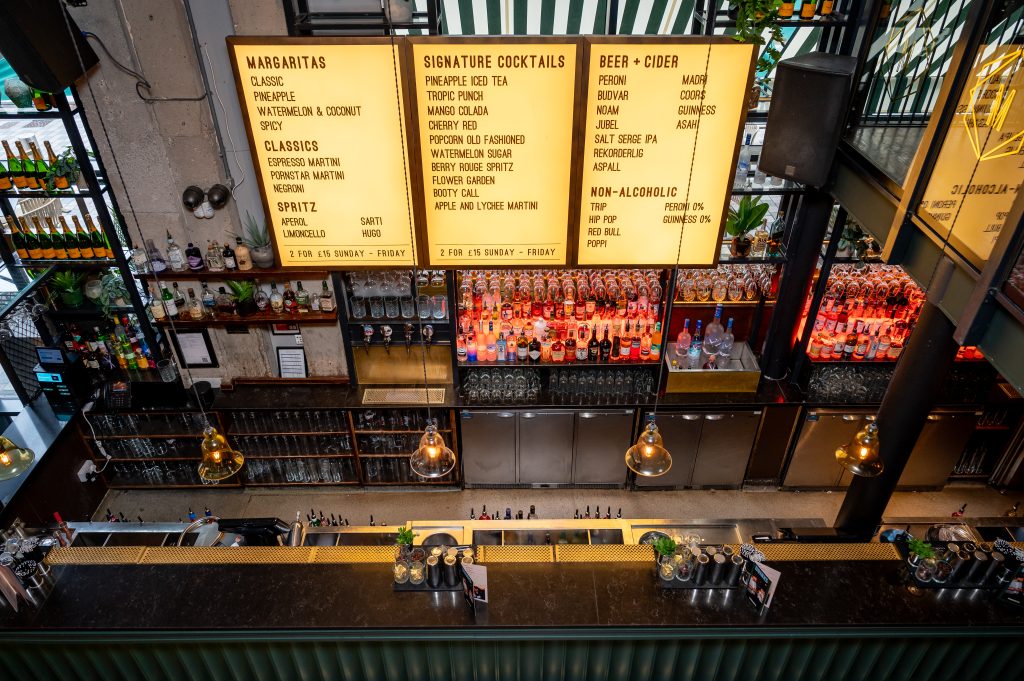 a well stocked bar with a large lightup menu board above it 